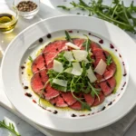 Vista aérea de un carpaccio de ternera servido en plato blanco, decorado con rúcula y lascas de parmesano.